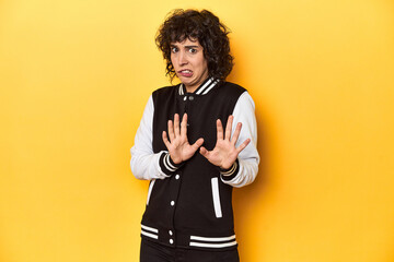 Curly-haired Caucasian woman in baseball jacket rejecting someone showing a gesture of disgust.