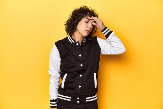 Curly-haired Caucasian woman in baseball jacket having a head ache, touching front of the face.