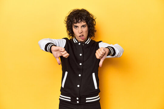 Curly-haired Caucasian woman in baseball jacket showing thumb down and expressing dislike.