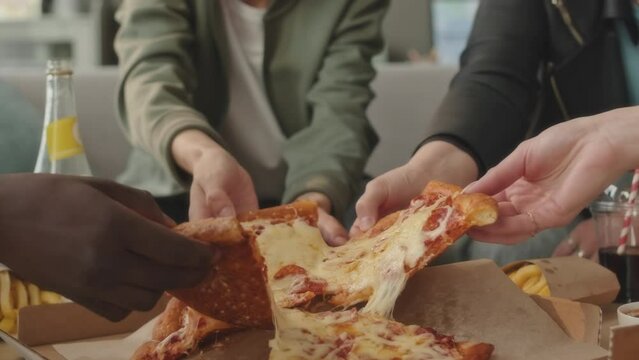 Unrecognizable multiethnic young people taking cheesy pizza slices from box while getting together in apartment to have good time