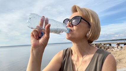 Portrait of elderly senior beautiful woman drinking water from plastic bottle at summer day in...
