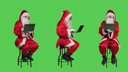 Modern santa character on laptop in full body greenscreen studio, sitting on chair and browsing...