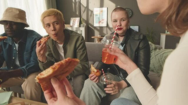 Handheld shot of company of young diverse gen z friends having good time together in bright apartment chatting, eating pizza and drinking soft drinks