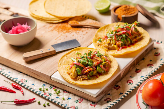 Tacos mexicanos de carnitas y tinga de pollo, sobre tabla de madera en una composici&oacute;n sobre mesa de restaurante grill con limones, tortillas, chiles y verduras con tajin