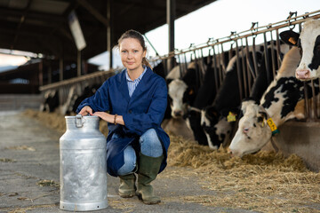 Positive female farmer with large metallic milk can in hangar with cows