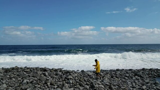 Man with a coffee wearing a yellow raincoat walking the Ruins of St. George, S&atilde;o Jorge beach at northeast coast