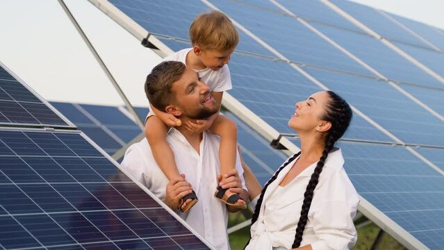 A young family of three is resting near a photovoltaic solar panels, smiling and kissing, concept of bright future