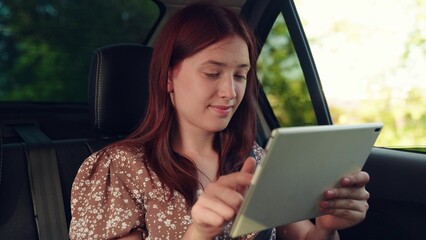 Young woman with tablet computer working remotely in back seat of car. Happy girl goes on journey. Student studies remotely. Businesswoman with digital tablet, car. Working online. Distance learning