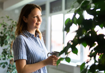 Portrait of positive woman with a mug in her hand among the plants next to the window in her house