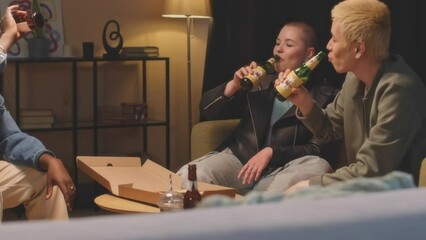 Medium shot of group of diverse friends clinking beer bottles and chatting while spending time together at house party at night