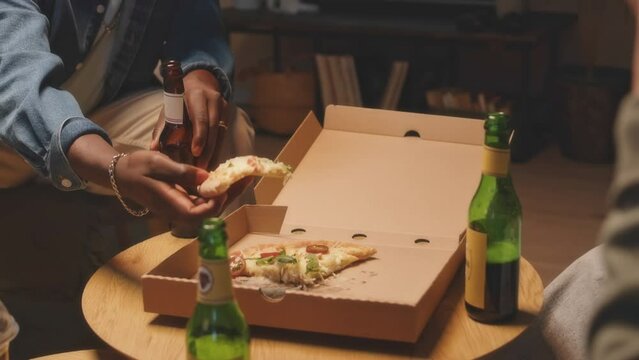 Medium shot of group of multiracial friends drinking beer and eating cheesy pizza while having good time together in apartment on weekend night