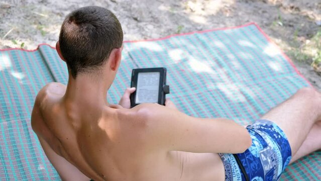 Male Reading an e-Book on a Colored Blanket while Relaxing in Beach. Back view. Close up. Head, shoulders. Nature. Smartphone in an old shabby case. Woods. Weekend. Blurred background. Outdoors.