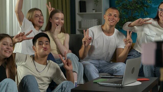 Medium shot of a group of teens, young people, friends sitting around the table, using a smartphone on a tripod to take a picture, photo together, showing peace gesture.