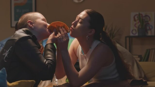 Medium shot of two cheerful girls taking bite on both sides of Mexican street taco while having fun during house party with friends on weekend
