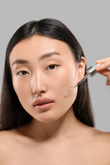 Fototapeta premium Beautiful young woman applying cosmetic serum onto her face on grey background