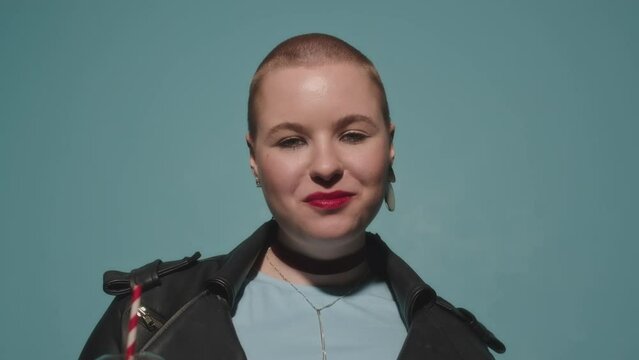 Tilt up of confident plus size Caucasian girl with shaved head taking bite of delicious pepperoni pizza and looking at camera standing on turquoise studio background wearing black leather jacket
