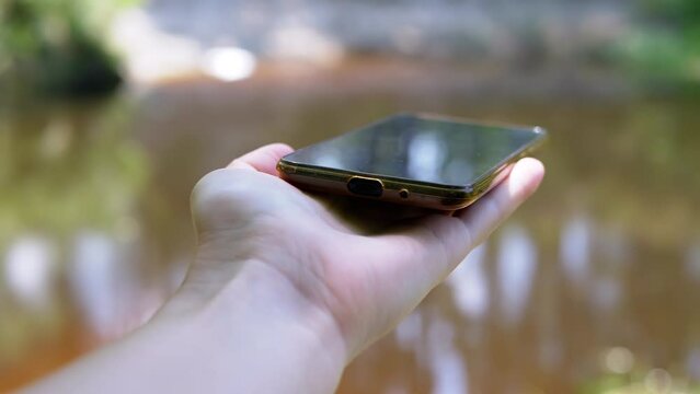 Searching for a Signal, Wi-Fi in the Forest using a Smartphone. A female hand holding out a mobile phone to blue sky, trying to catch a signal. Bad or no signal. Blurred water background. Close up.