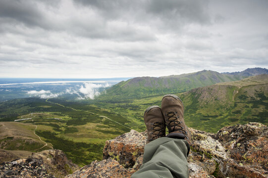 Pov Of Hikers Legs And Feet As He Rests And Enjoys The View From Flat Top Mountain Overlooking The Anchorage Bowl, Southcentral Alaska, Summer