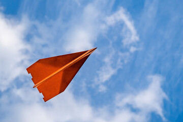 Orangefarbener fliegender Papierflieger von unten zu sehen, im Hintergrund leicht bewölkter blauer...