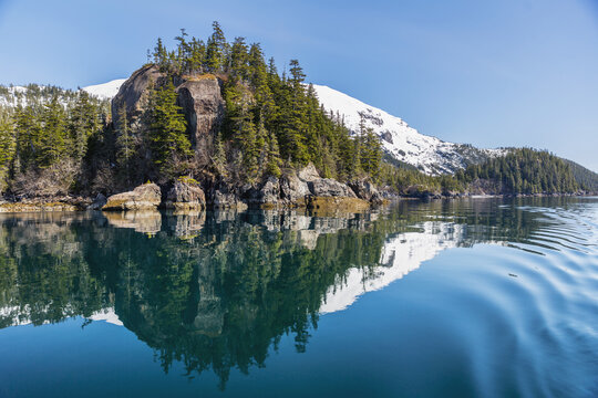 A Tall Outcropping Of Rocks And Evergreen Trees Reflects On The Waters Of Prince William Sound; Whittier, Alaska, United States Of America