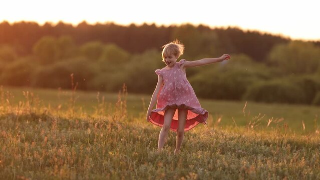 A happy young girl in a summer dresss spins on the field against the backdrop of the evening sun and throws her hat up. Carefree holiday concept. Front view.