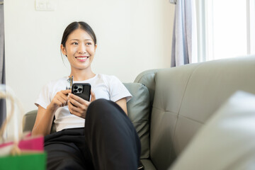 Asian woman holding credit card and smart phone and laptop sitting on sofa at home doing online banking transactions in online shopping