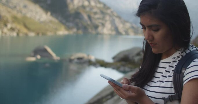 Young female trekker using her phone to check the maps by a mountain lake in the Swiss alps and looking at the camera