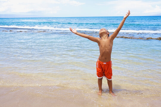 A young boy standing in the shallow water of the ocean with arms raised and looking up; Hawaii united states of america