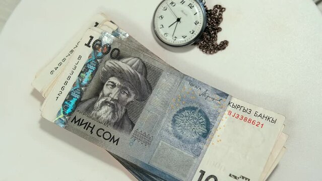 Stack 1000 som bills and vintage pocket watch on chain rotate on white surface, money background. Close-up one thousands som national currency Republic of Kyrgyzstan