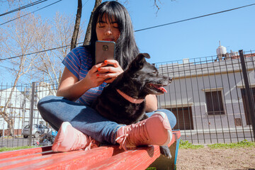 young latin woman with her dog in park watching social networks on her phone