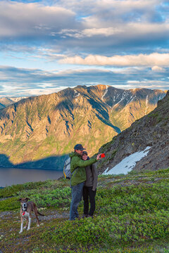 Couple taking a picture of themselves on top of a mountain at twilight with beautiful scenic backgound, while their dog stands beside them looking forward; Carcross, Yukon Territory, Canada