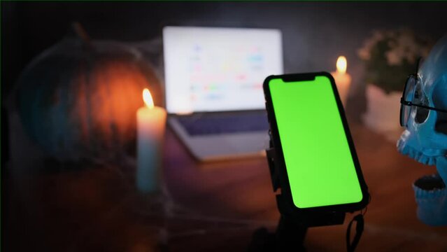 Creepy skeleton enthusiastically scrolls screen smartphone with chromakey among cobwebs and burning candles at desktop with pumpkin and  open laptop. Halloween atmosphere. Cyberstalking and disguise.