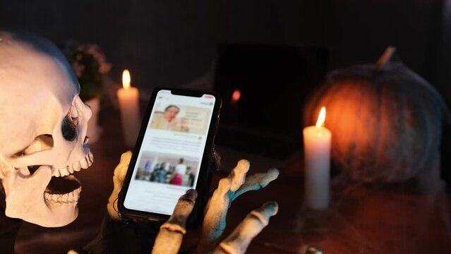 Side view close up death character halloween costume uses phone, scrolling feed, likes, sitting dark room in fog, pumpkin and candles in cobwebs. Cyberstalking and disguise. Halloween party invitation