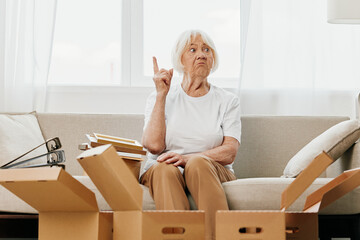Fototapeta na wymiar elderly woman sits on a sofa at home with boxes. collecting things with memories albums with photos and photo frames moving to a new place cleaning things and a happy smile. Lifestyle retirement.