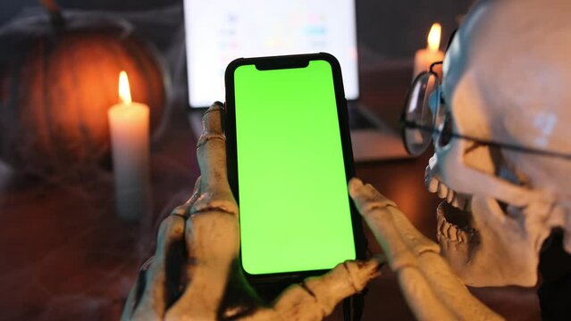 Сreepy skeleton holds phone with chromakey in bony hands and feverishly scrolls smartphone screen in fog by candlelight next to laptop and pumpkin in cobweb in honor of Happy Halloween. Rear view shot