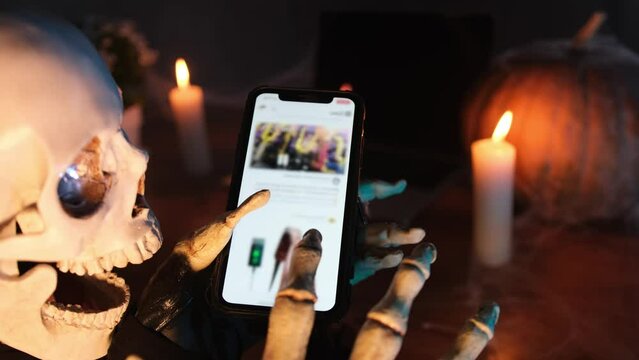 In swirling fog mysterious character in skeleton costume in image of death locks screen smartphone, scrolls through feed on social network, examines photo in light candles, close-up. Halloween party.