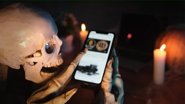 In dark crypt puffs of smoke, skeleton uses modern smartphone scrolls through social networks and watches photos and online magazine, with lit candles and pumpkin. Happy Halloween.