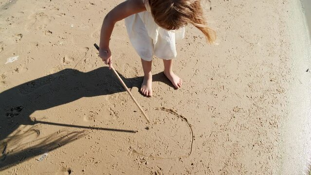 Girl child drawing with a stick on the sand on the beach and having fun