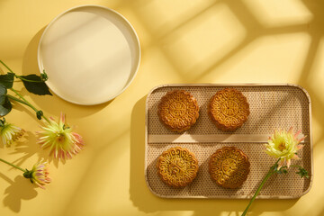 Fototapeta na wymiar Scene for advertising mooncake product on yellow background with window shadow. On bamboo tray, mooncakes are displayed, decorated with flowers and empty plate for product presentation