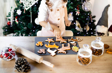 Happy little girl coloring Christmas gingerbread cookies at home. Christmas decoration in the kitchen. Fir tree with fairy lights. The concept of the New Year and Christmas