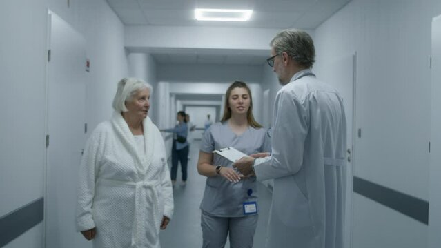 Mature doctor walks the clinic corridor, comes to colleague and elderly patient. Physician shows test results on papers to nurse and woman. Medical staff and patients in hospital hallway. Slow motion.