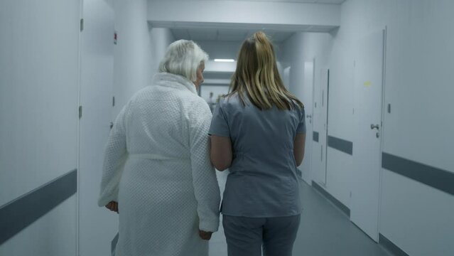 Back view of female medic with digital tablet and elderly woman talking, walking along the clinic corridor to hospital room. Medical staff, doctors and patients in medical center hallway. Slow motion.