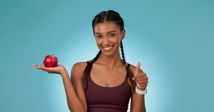 Woman, athlete and apple in hands with thumbs up, yes and healthy decision for nutrition, wellness and benefits in diet. Happy, nutritionist and choice of fruits or food on blue background in studio