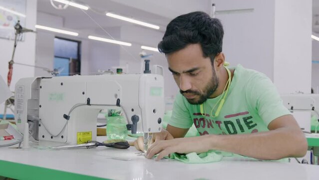 A skilled Indian young man or male artisan or tailor is working on a sewing machine along with other workers in a busy cotton or textile workshop or garment industry or factory.