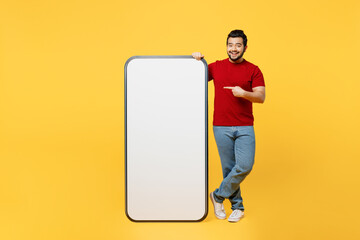 Full body young Indian man wears red t-shirt casual clothes point finger on big huge blank screen mobile cell phone smartphone with area isolated on plain yellow orange background. Lifestyle concept. © ViDi Studio