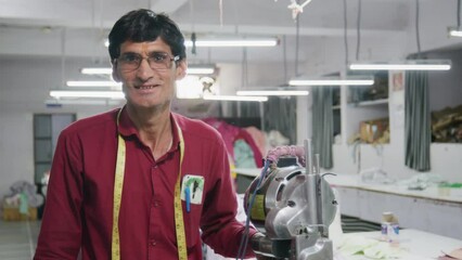 A Skilled happy Indian man or male tailor is standing and facing the camera smiling next to the specialized machine in an indoor textile workshop or a small scale garment factory or industry
