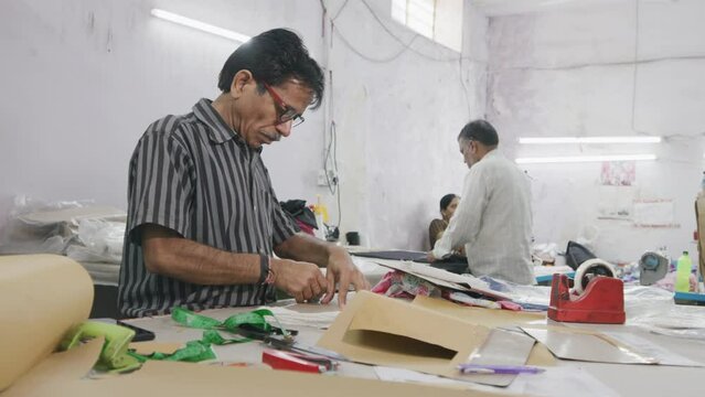 A middle aged Indian man or male skilled tailor or artisan in standing and busy working on the cloth or cotton garment designs in a textile workshop or small business or factory.