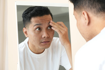 Obraz premium Photo of anxious Asian young man looking in the mirror worried about wrinkles or acne on unhealthy skin.