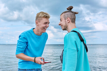 Two friends cheerful laughing young guys with smartphone, beach outdoor