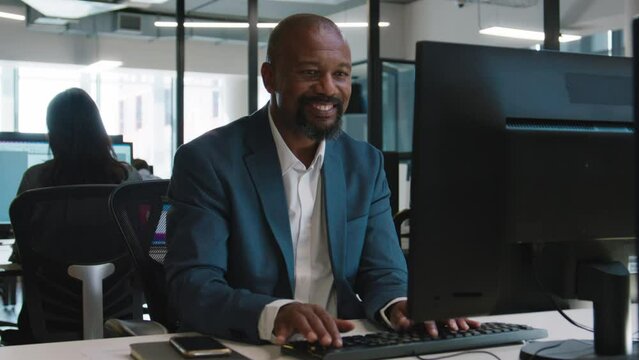Happy mature black man in businesswear typing on computer at desk in office